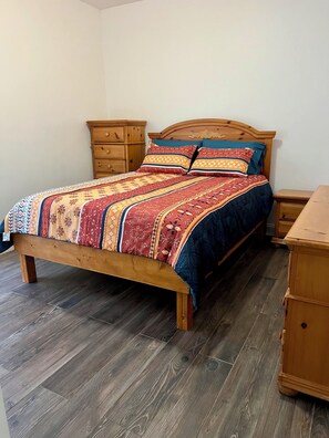 Ein hölzernes Bett mit gemustertem Bettbezug und Kissen, ein Nachttisch aus Holz sowie eine Holzkommode in einem Zimmer mit Holzboden.