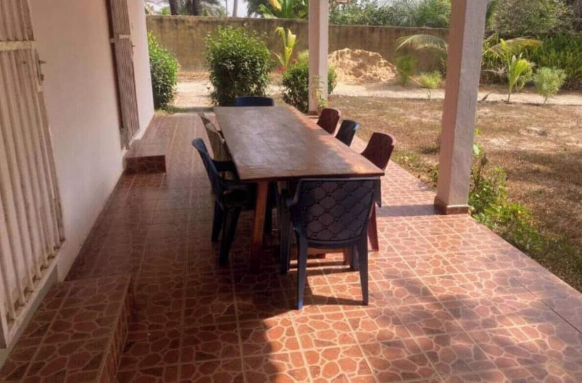 Een overdekt terras met een rechthoekige tafel en stoelen, omgeven door een betegelde vloer.