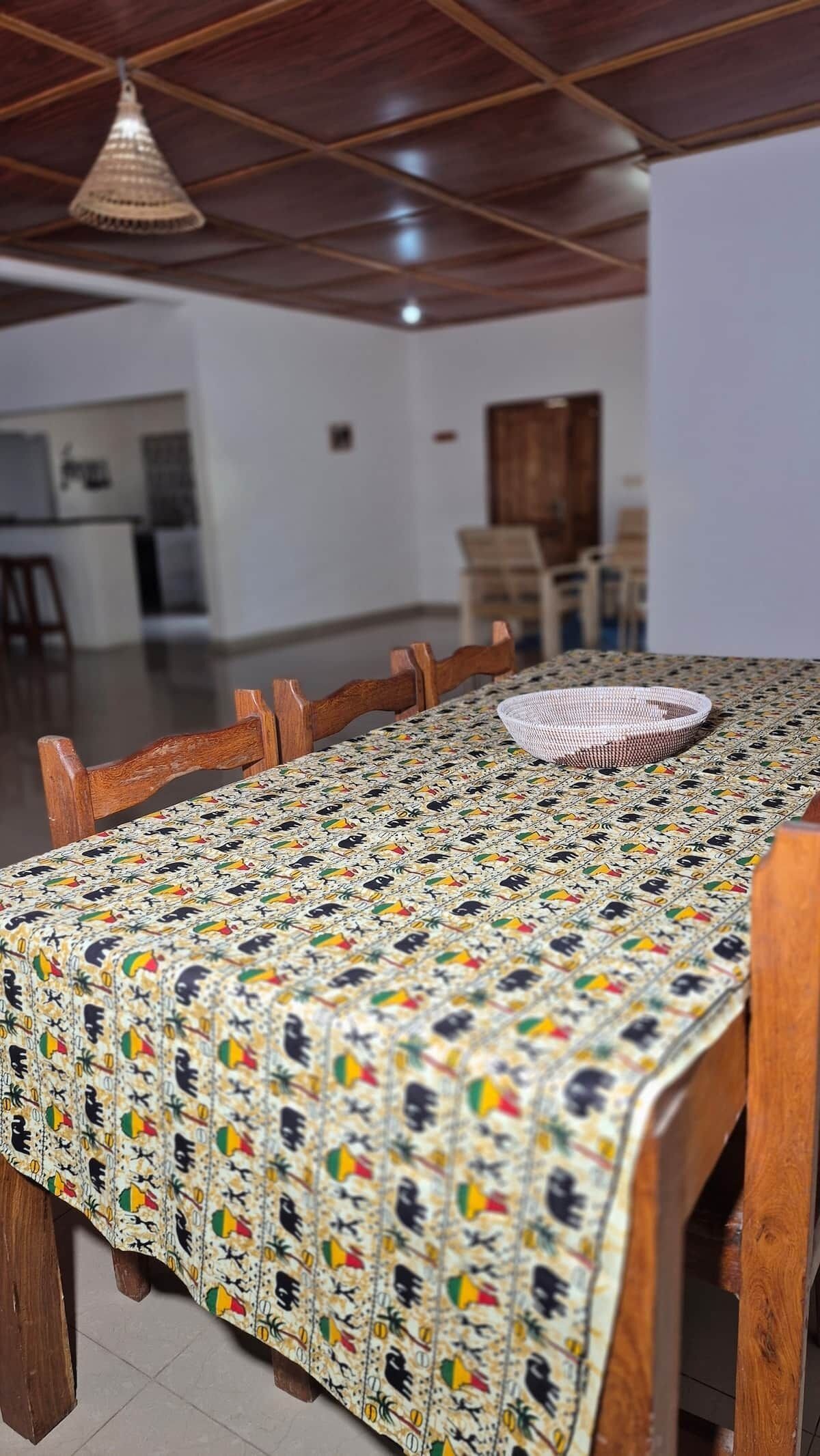 Ein Essbereich mit einem langen Holztisch, bedeckt mit einer gemusterten Tischdecke, umgeben von Holzstühlen.