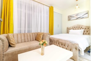 Quarto de hotel com sofá bege, mesa de centro branca, cama com cabeceira estofada e cortinas amarelas.