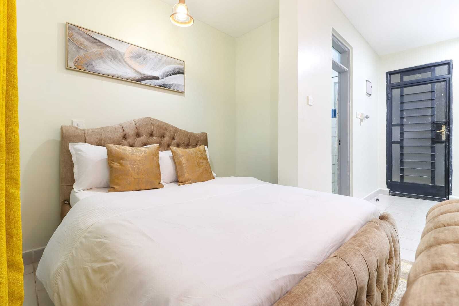 A neatly made bed with a tufted headboard and multiple pillows, a framed abstract artwork above it, and a door with a black shutter to the right.