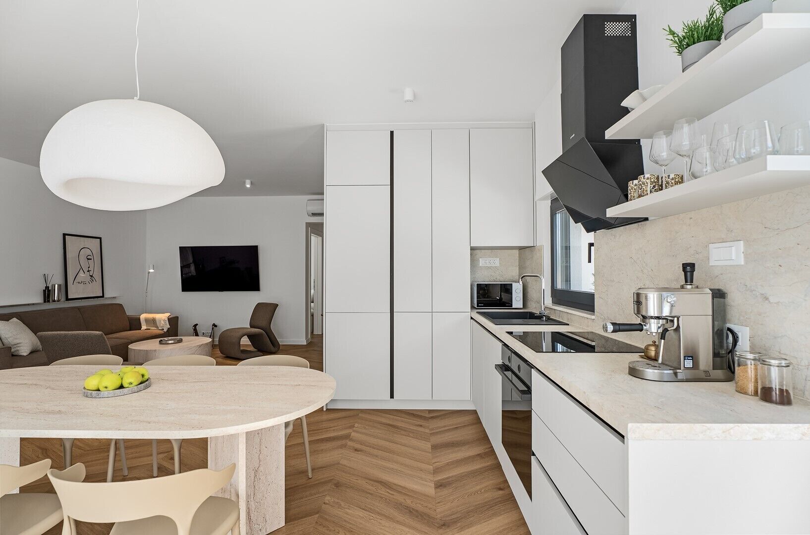A modern kitchen with white cabinetry, a round dining table with chairs, a coffee machine, and a built-in oven.