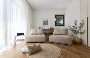 Ein modernes Wohnzimmer mit einer Sofagarnitur, einem runden gewebten Teppich, einem Beistelltisch, einer Lampe, einem Bücherregal und einem Blumentopf.