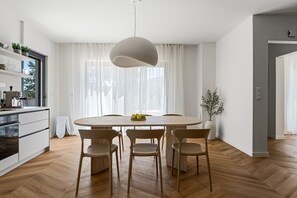 Een moderne keuken met een eethoek, met een ronde tafel met stoelen, een hanglamp en een groot raam met lichte gordijnen.