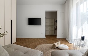 Een moderne woonkamer met een flatscreen televisie aan de wand, een houten visgraatvloer en een groot raam met dunne gordijnen.