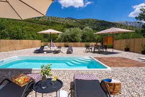 A swimming pool with lounge chairs and umbrellas, surrounded by a pebble-covered area with a wooden deck and a view of a mountainous landscape. - Villa Three Olives: A Perfect Family Retreat near Omis (Naklice)