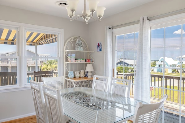 Una habitación iluminada por el sol, con una mesa de vidrio y sillas de mimbre blanco, un estante con objetos decorativos, y vistas a una zona residencial.