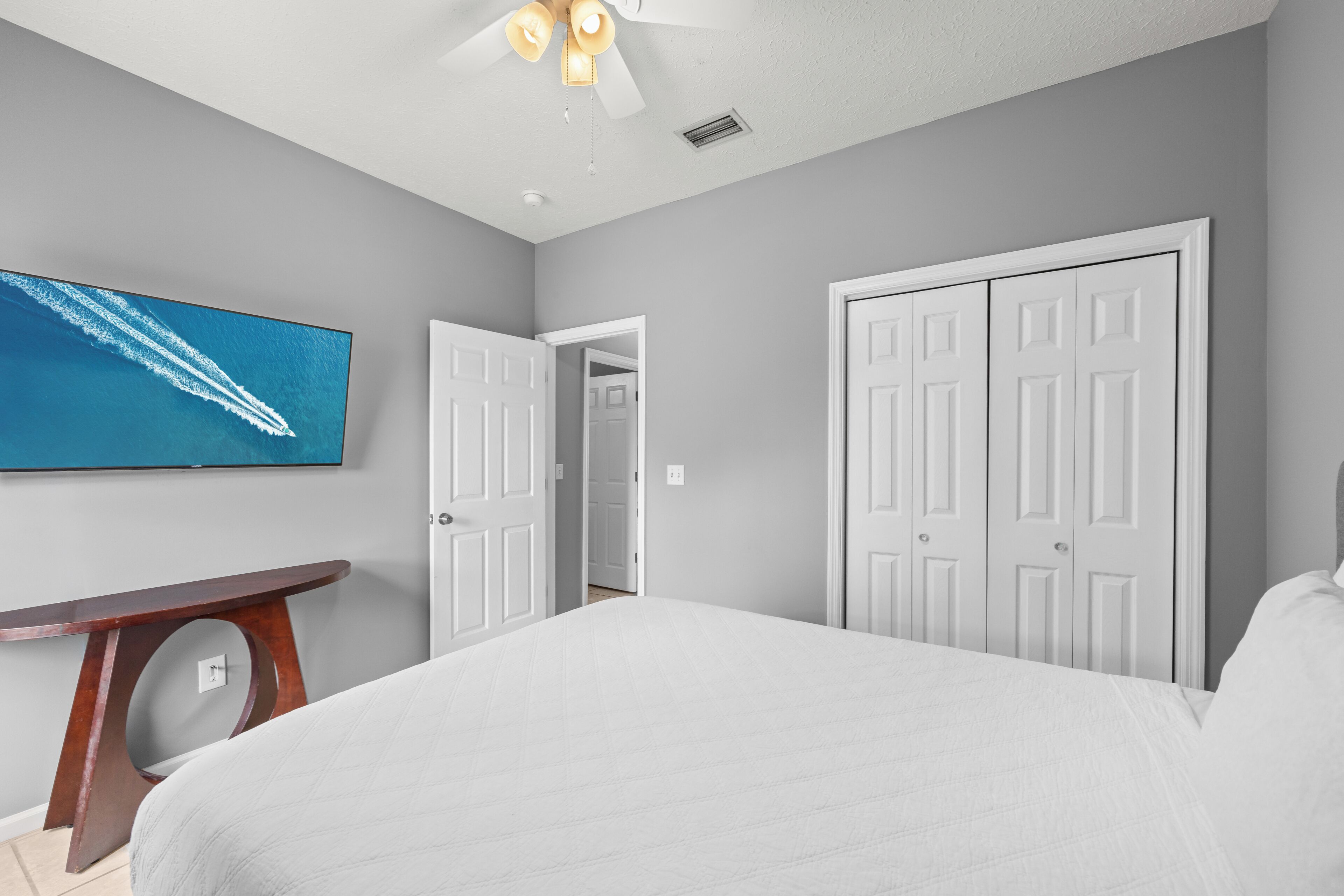 A bedroom with a bed, a TV, a ceiling fan, and a door leading to another room.