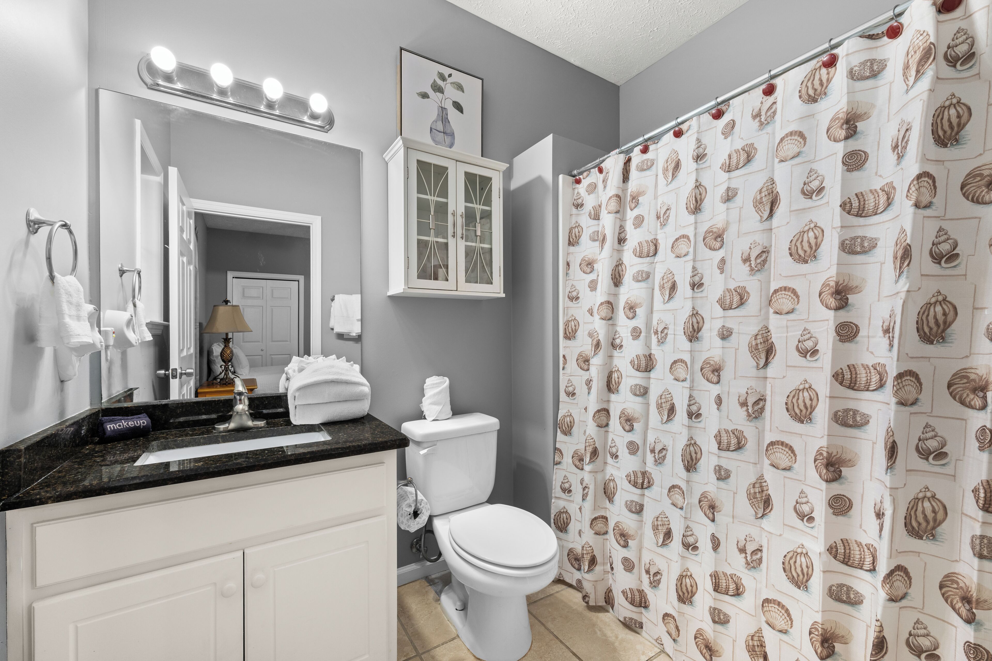 Een badkamer met een wastafel, toilet, douchegordijn met schelpmotief, en een spiegel met verlichting erboven.