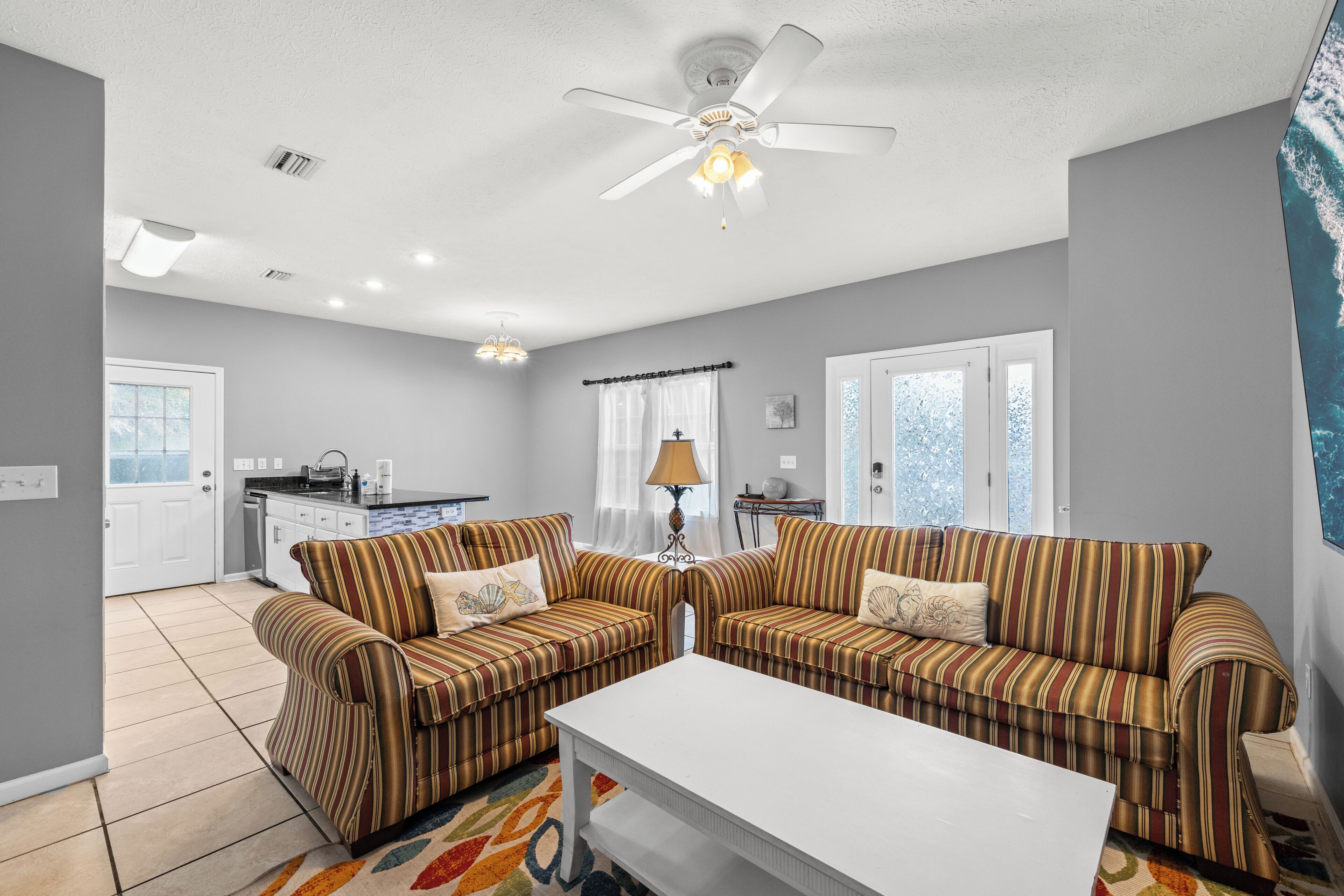 A living room with striped sofas, a white coffee table, a ceiling fan, and a kitchen in the background.