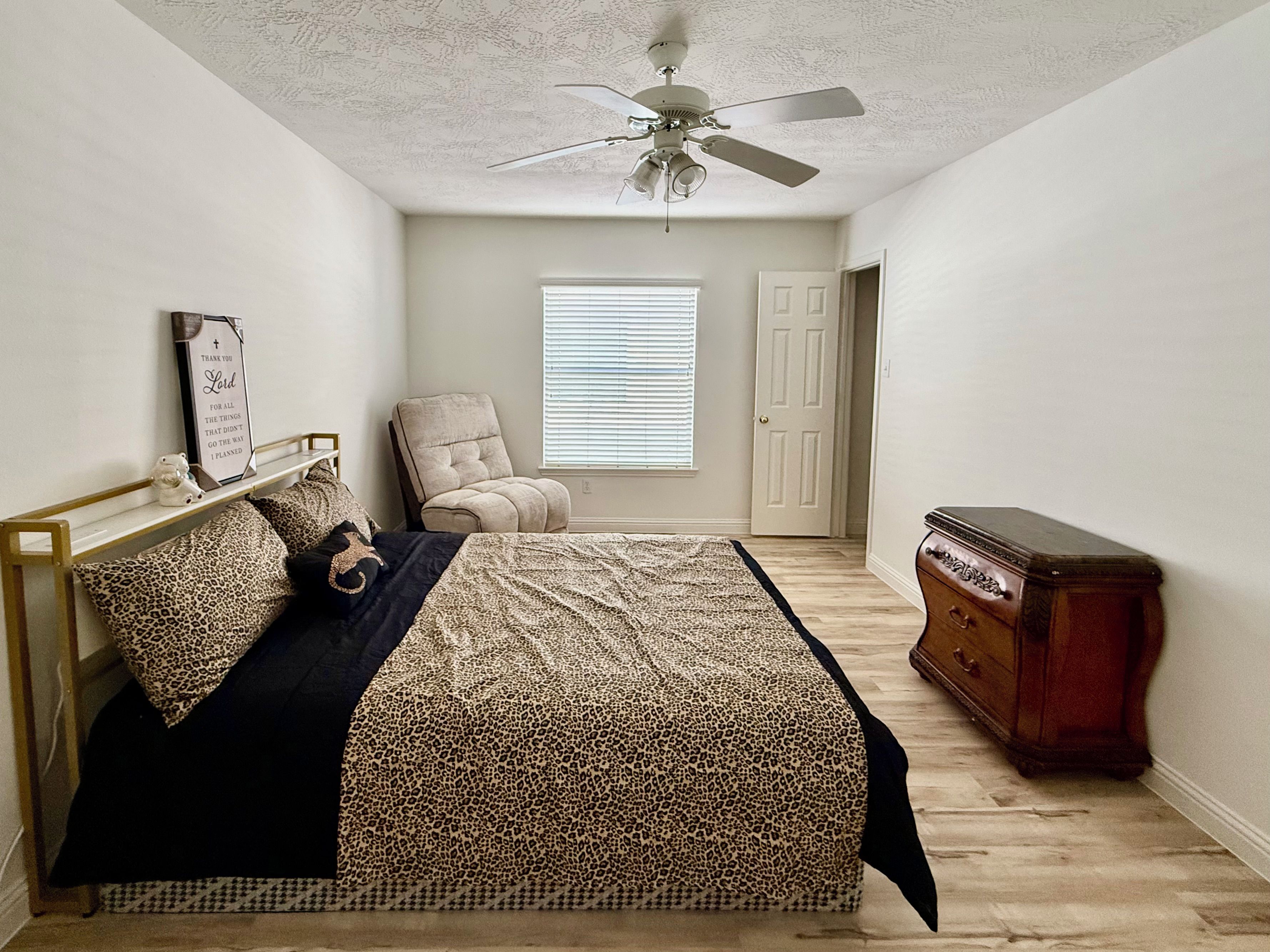 Une chambre à coucher comprenant un lit, une chaise, une commode, un ventilateur de plafond et une fenêtre avec des stores.