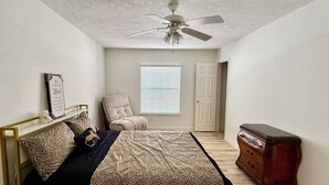 Ein Schlafzimmer mit einem Bett, einem Sessel, einer Kommode, einem Deckenventilator und einem Fenster mit Jalousien.