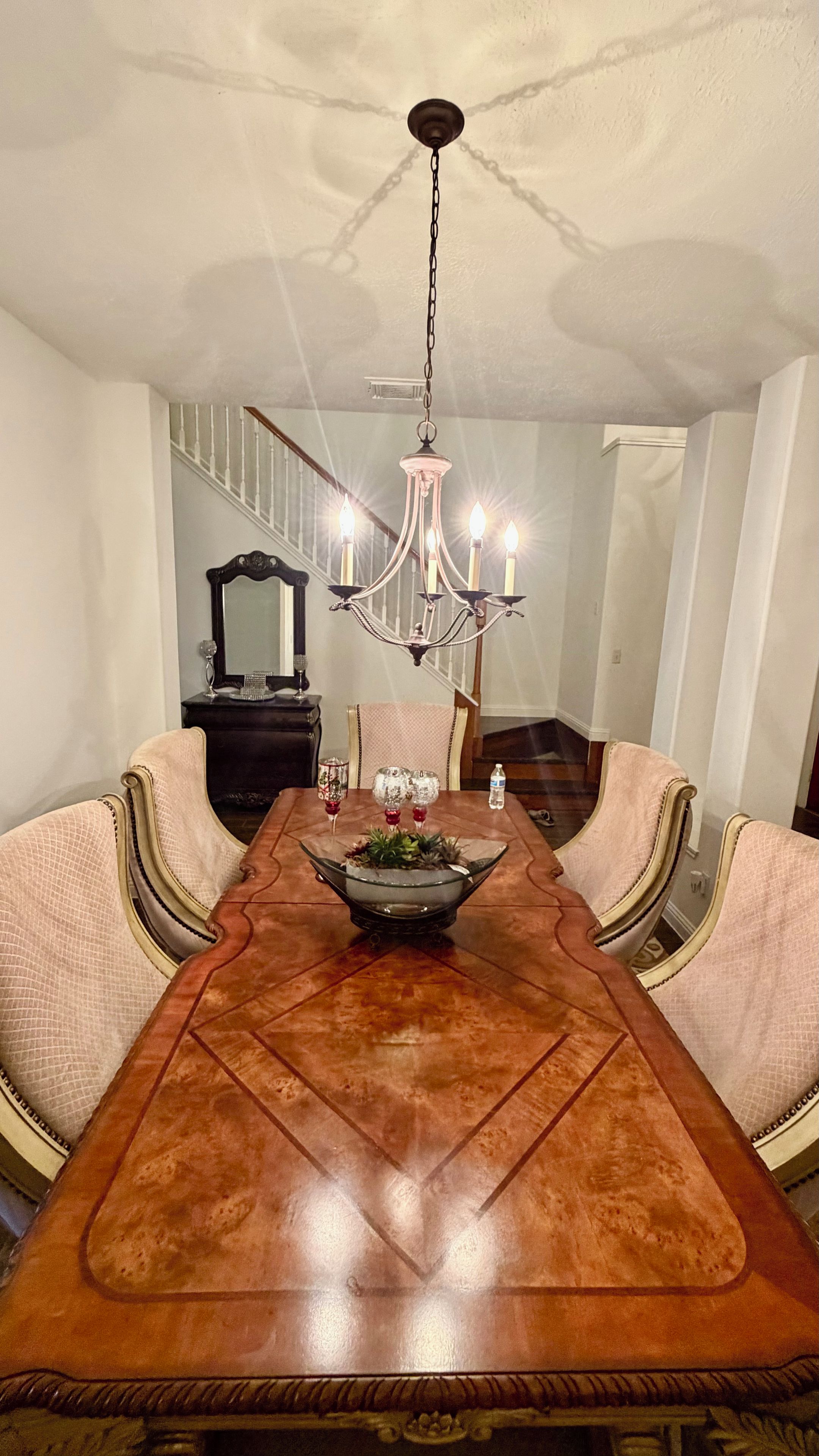A dining room with a long wooden table, cushioned chairs, a chandelier, and a staircase in the background.