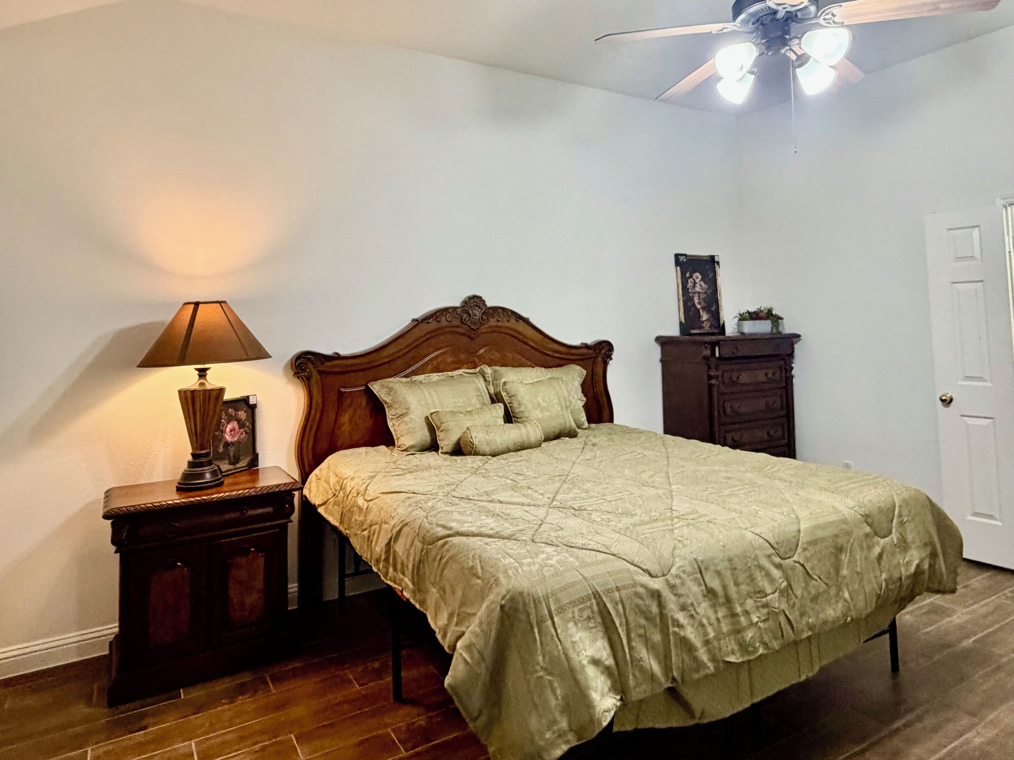 Une chambre à coucher comprenant un lit, une table de chevet, une lampe, une commode et un ventilateur de plafond.
