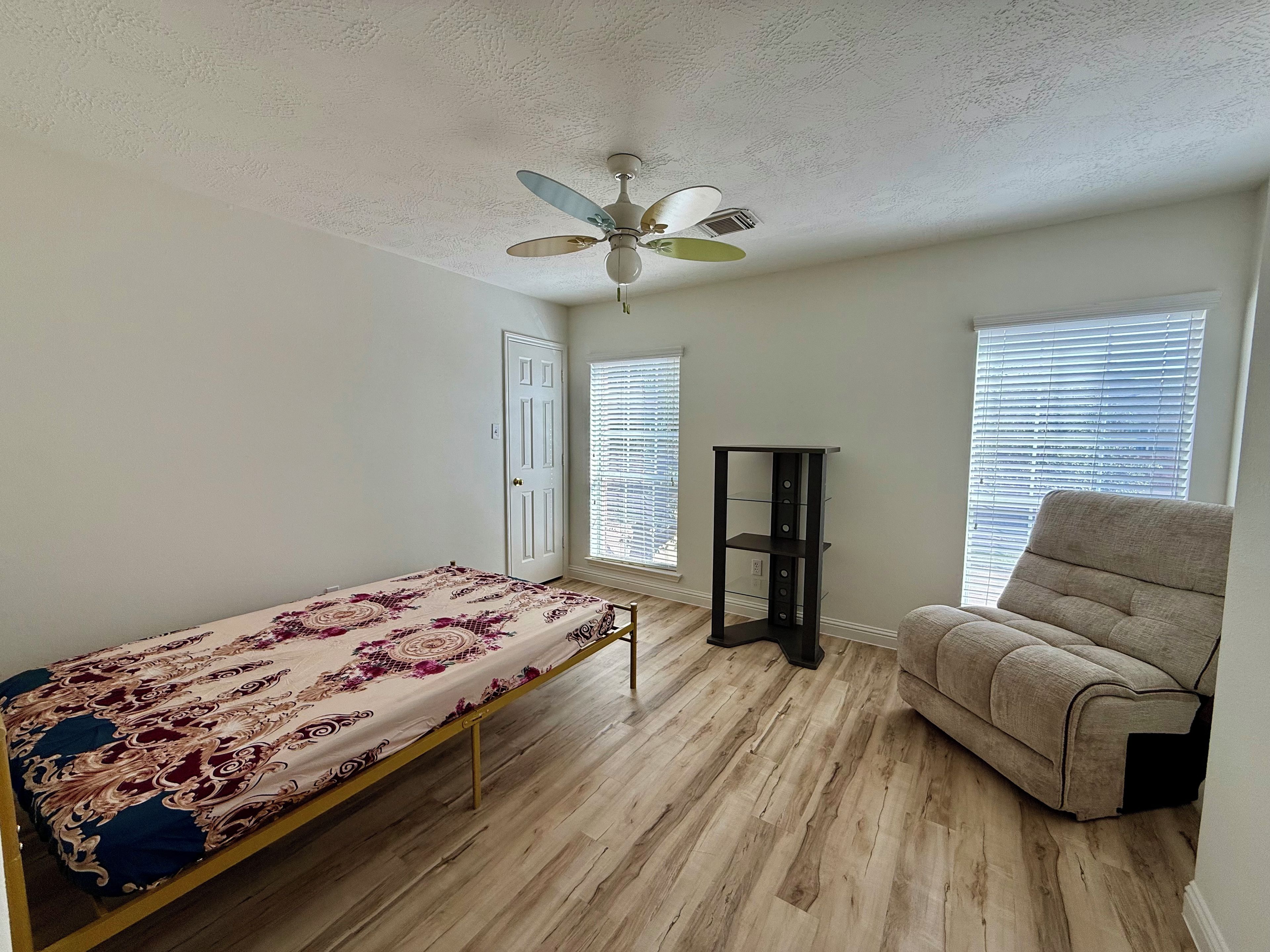 Une pièce comprenant un lit, une chaise, un bureau debout et un ventilateur de plafond.