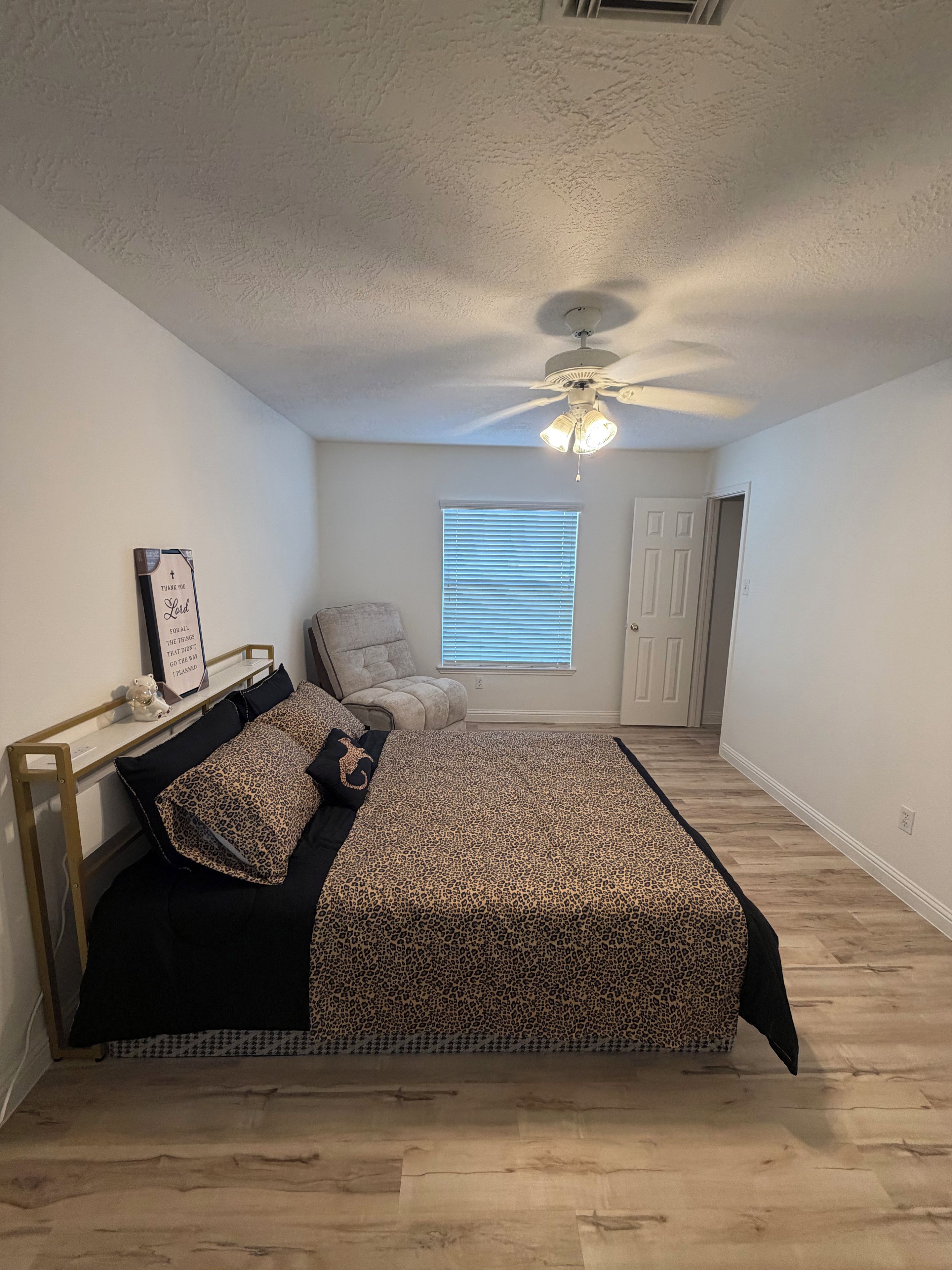 A bedroom with a bed, a chair, a window with blinds, a ceiling fan, and a door.