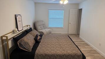 A bedroom with a bed, a chair, a window with blinds, a ceiling fan, and a door.
