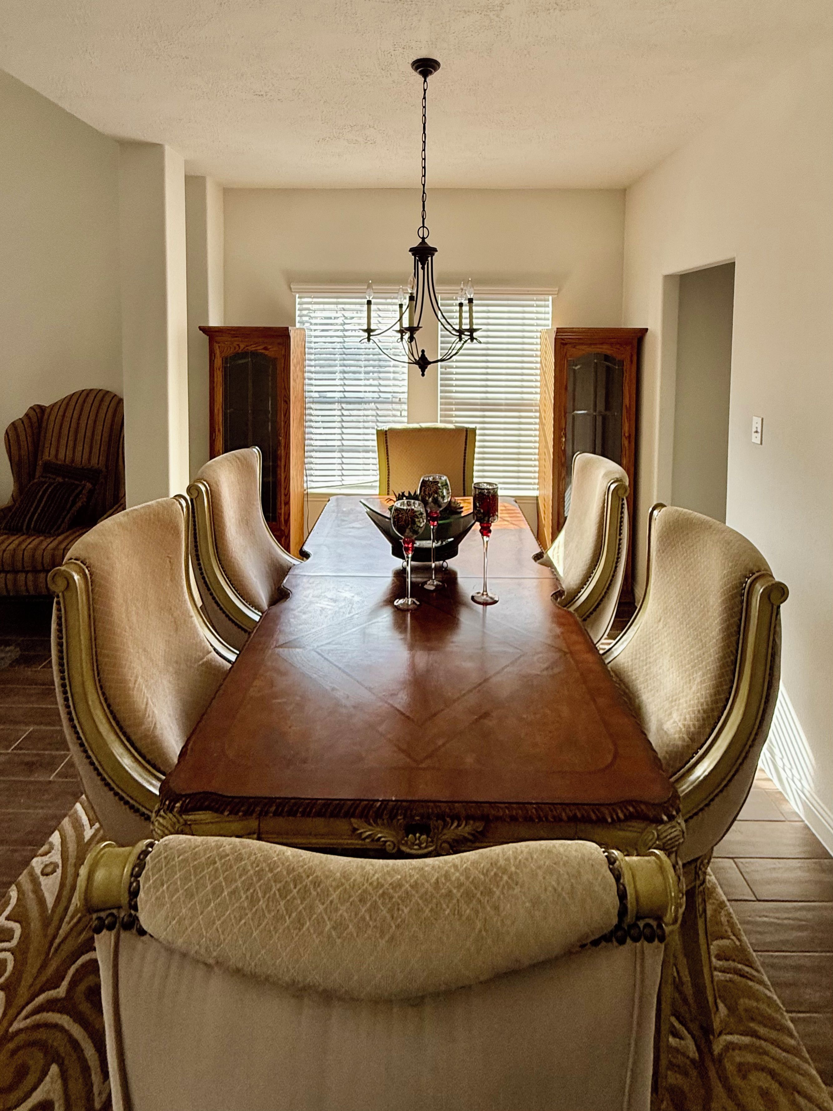 Une salle à manger avec une longue table en bois, six chaises rembourrées, un lustre et une fenêtre avec des stores.