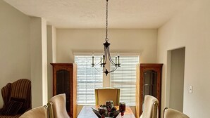 Ein Esszimmer mit einem langen Holztisch, sechs gepolsterten Stühlen, einer Chandelierleuchtre und einem Fenster mit Jalousien.