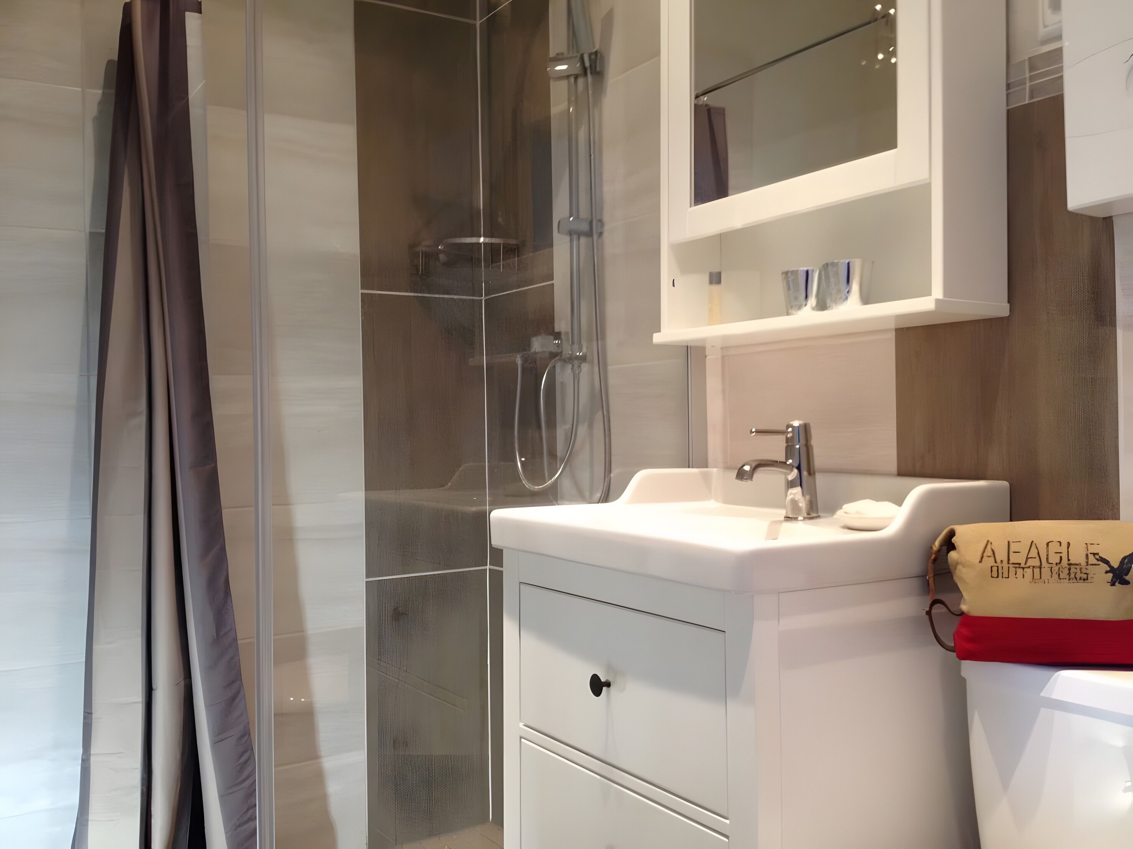Une salle de bain moderne avec une douche à parois de verre, un lavabo blanc avec un miroir au-dessus, et des toilettes sur lesquelles est posé un sac beige.