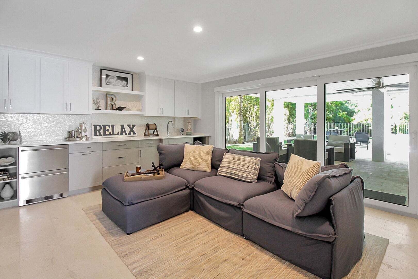 A modern kitchen with white cabinets, a grey sectional sofa, and a large sliding glass door leading to an outdoor patio.