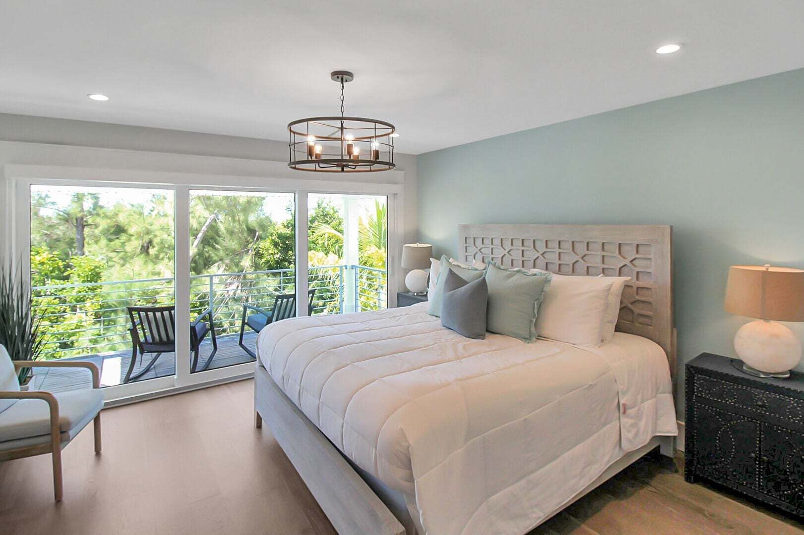 A bedroom with a large bed, a nightstand, a chair, and a balcony with a view of greenery.