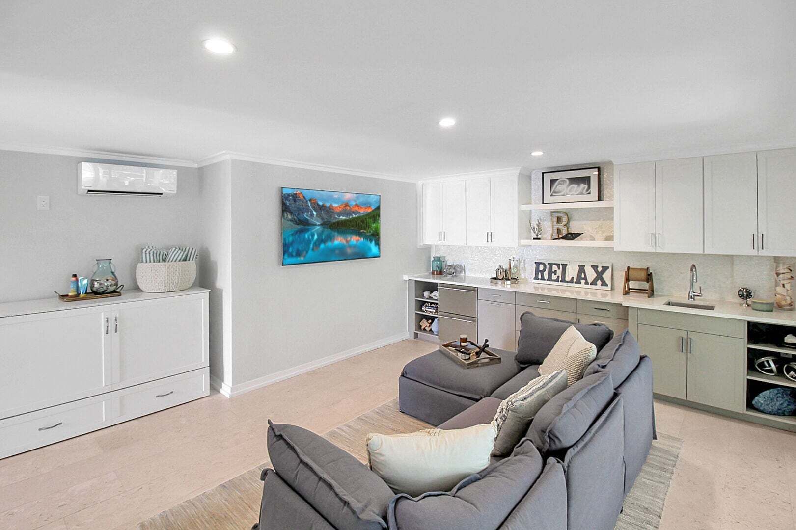 A modern living room with a grey sectional sofa, a kitchen with white cabinets, and a painting of a mountain landscape.