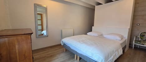 A bedroom with a wooden floor, a bed with white bedding, a wooden dresser, a radiator, and a mirror on the wall.