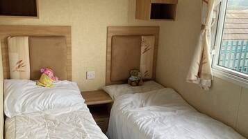 A room with two beds, each with a headboard and a bedside table. There is a window with curtains, and a view of the outside.