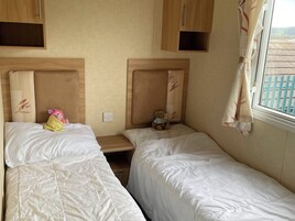 A room with two beds, each with a headboard and a bedside table. There is a window with curtains, and a view of the outside.