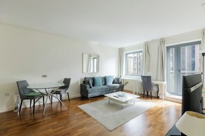 Una sala de estar moderna con una mesa de comedor de vidrio, un sofá gris y un amplio ventanal con cortinas.