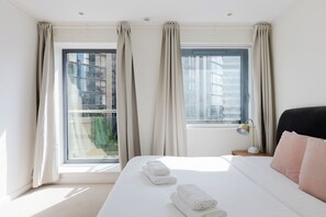 A hotel room with a large bed, two windows with curtains, a bedside lamp, and a view of buildings outside. - Sleek 1BDR City Stay In Canary Wharf (Greater London)