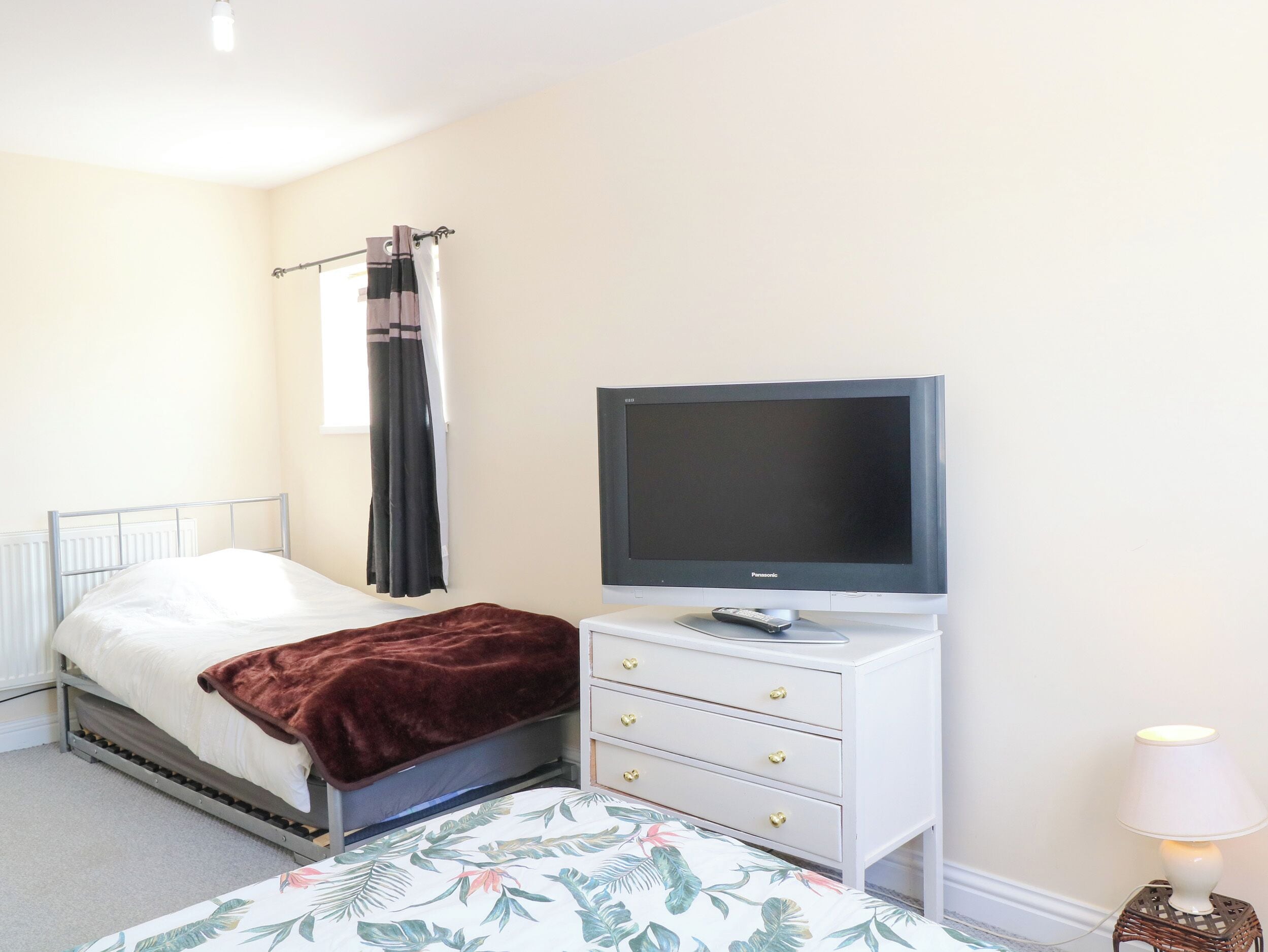 Une chambre à coucher avec un lit, un téléviseur posé sur un meuble, une commode avec des tiroirs, une lampe et une fenêtre avec des rideaux.