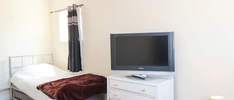 Une chambre à coucher avec un lit, un téléviseur posé sur un meuble, une commode avec des tiroirs, une lampe et une fenêtre avec des rideaux.