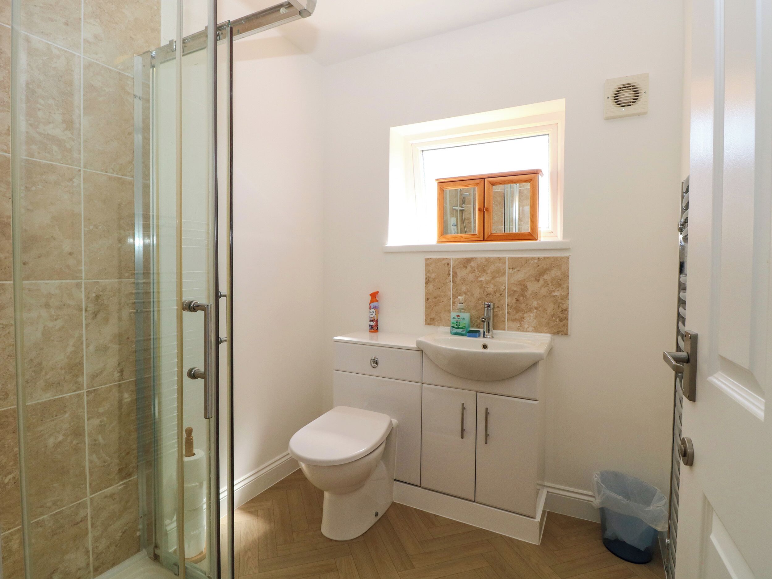 Ein Badezimmer mit Duschkabine aus Glas, Toilette, Waschbecken und einem Fenster darüber sowie einem blauen Mülleimer.