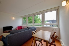 Ein Zimmer mit einer Couch, einem Tisch, einem Fernseher und großen Fenstern mit Blick auf Bäume und Gebäude.