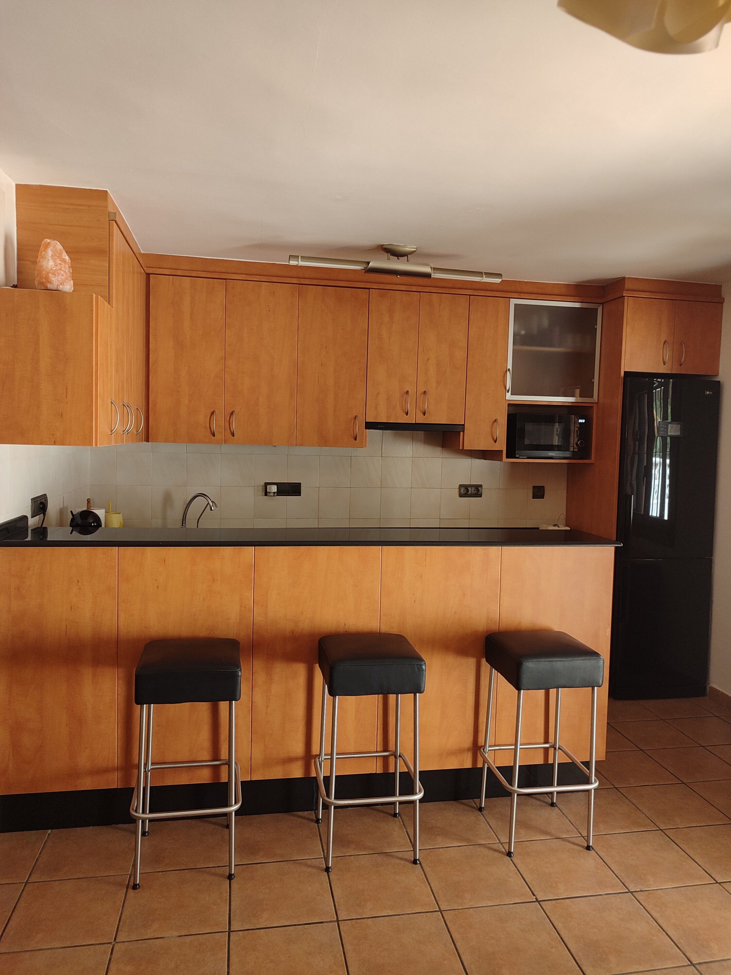 A kitchen with wooden cabinets, a black refrigerator, a microwave, and three black bar stools.