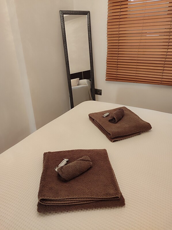 A neatly made bed with two brown towels and a pillow, a large mirror, and a window with blinds.
