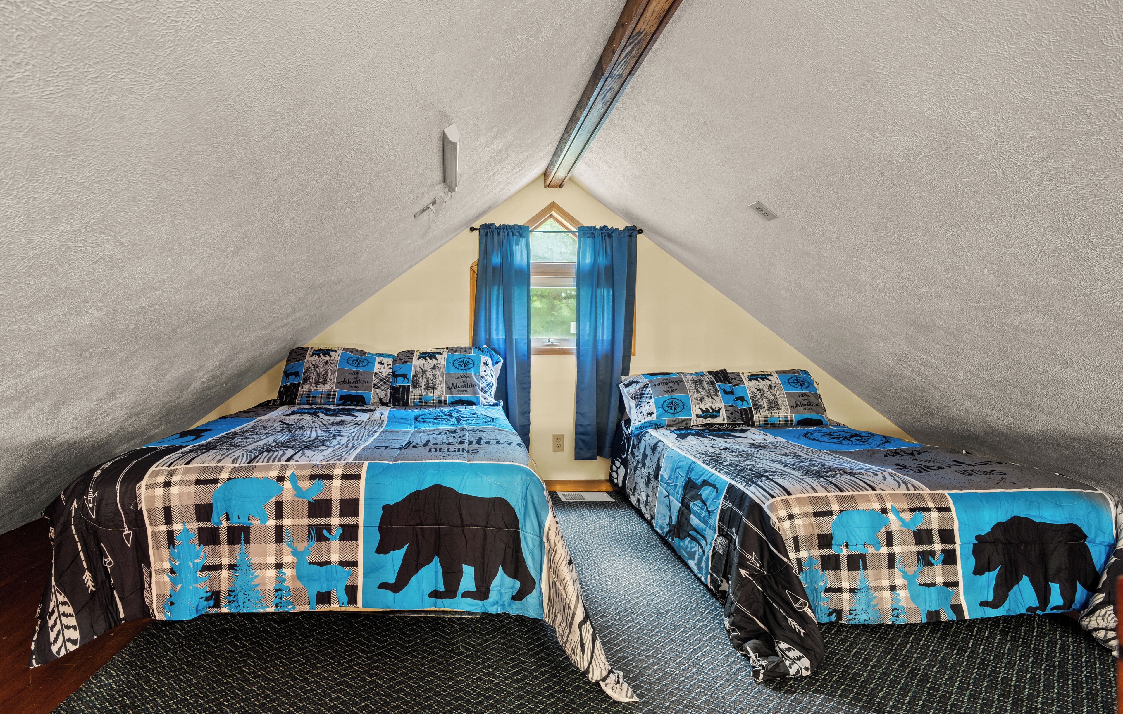 A triangular attic bedroom with two beds, each with a bear-themed bedspread, a window with blue curtains, and a sloped ceiling.