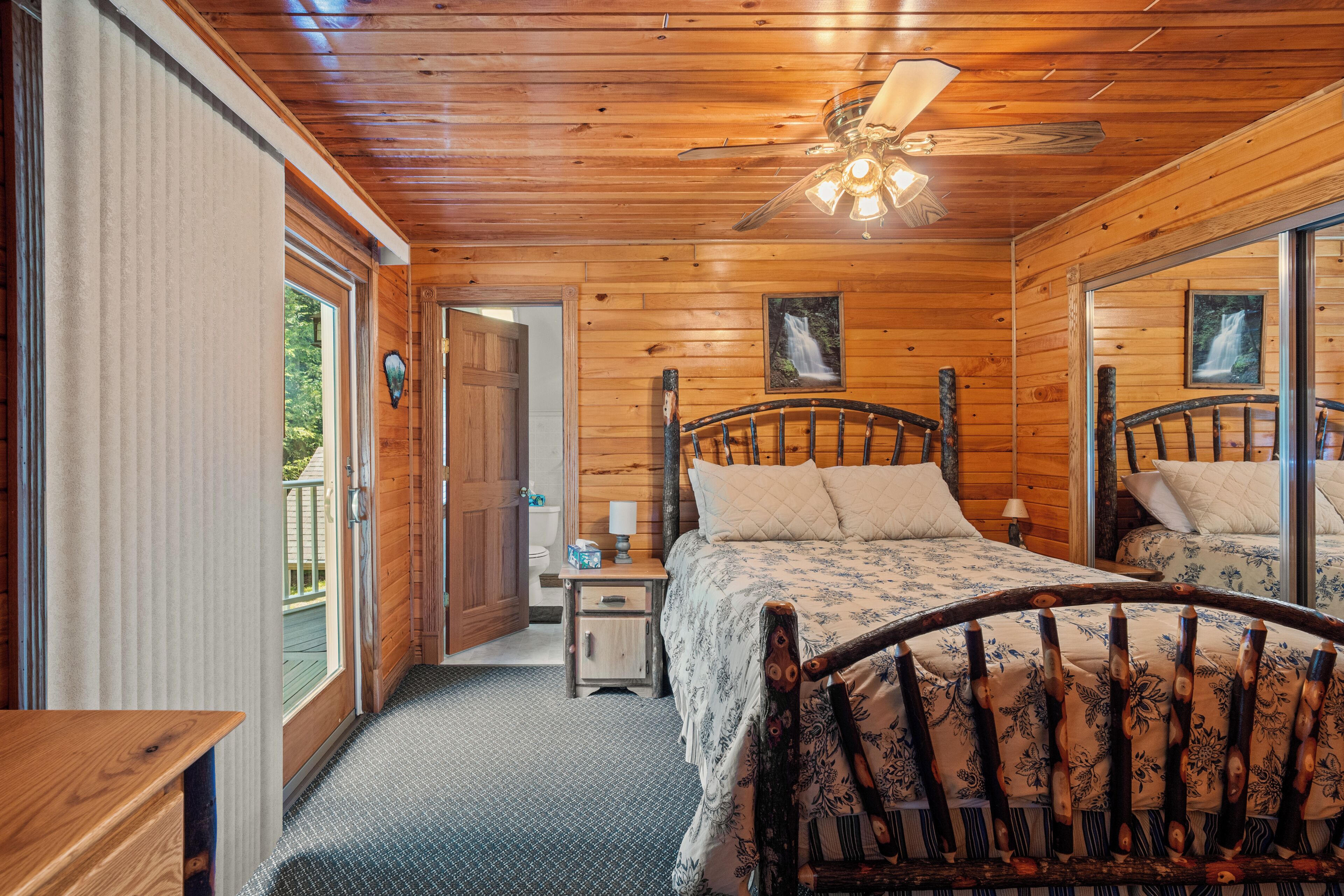 Une chambre dans une cabane en bois, avec un lit, des tables de chevet, un ventilateur de plafond et une vue sur l’extérieur.