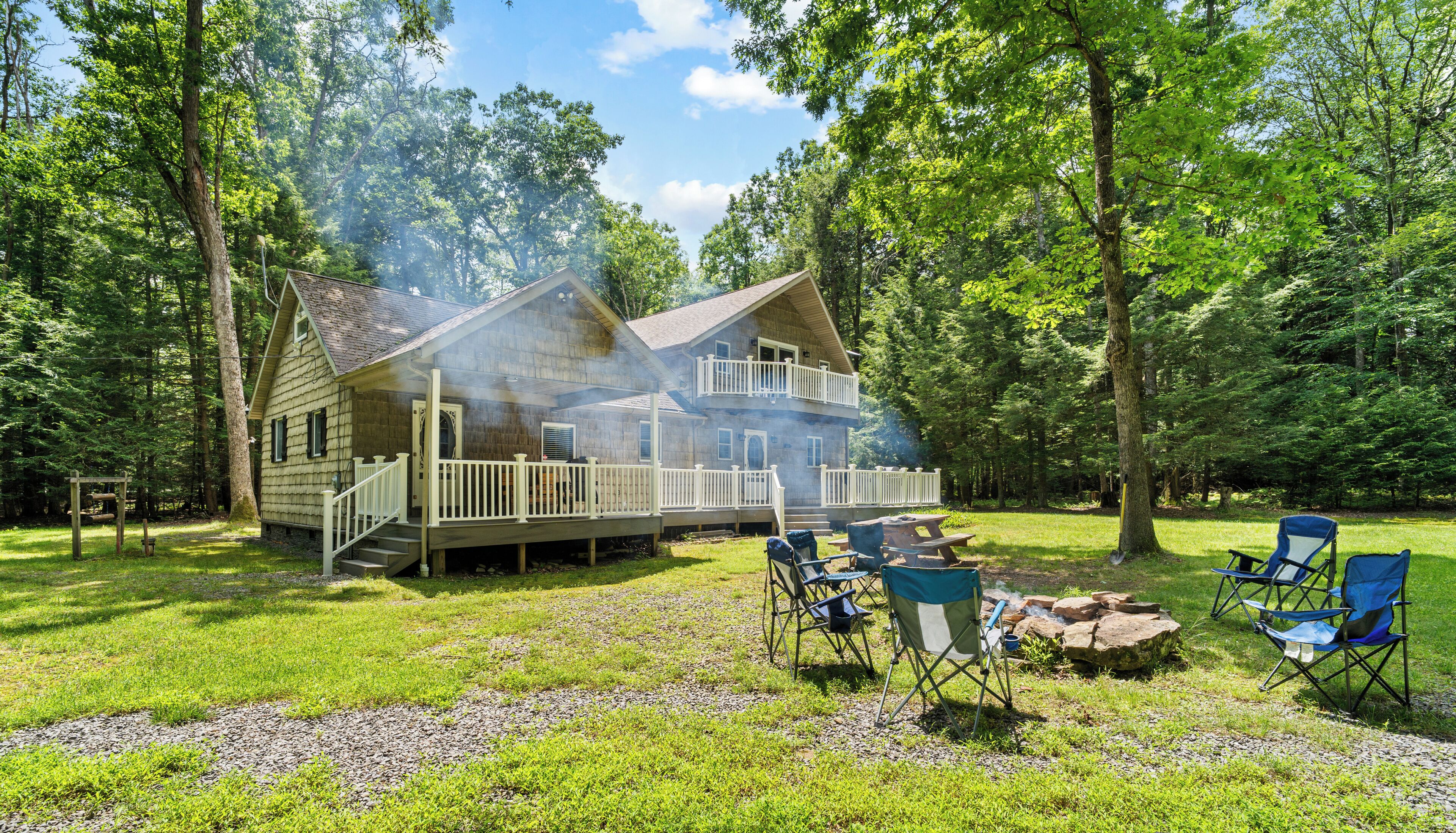 Une cabane à deux étages avec une terrasse, entourée d’arbres, et un foyer avec des chaises.