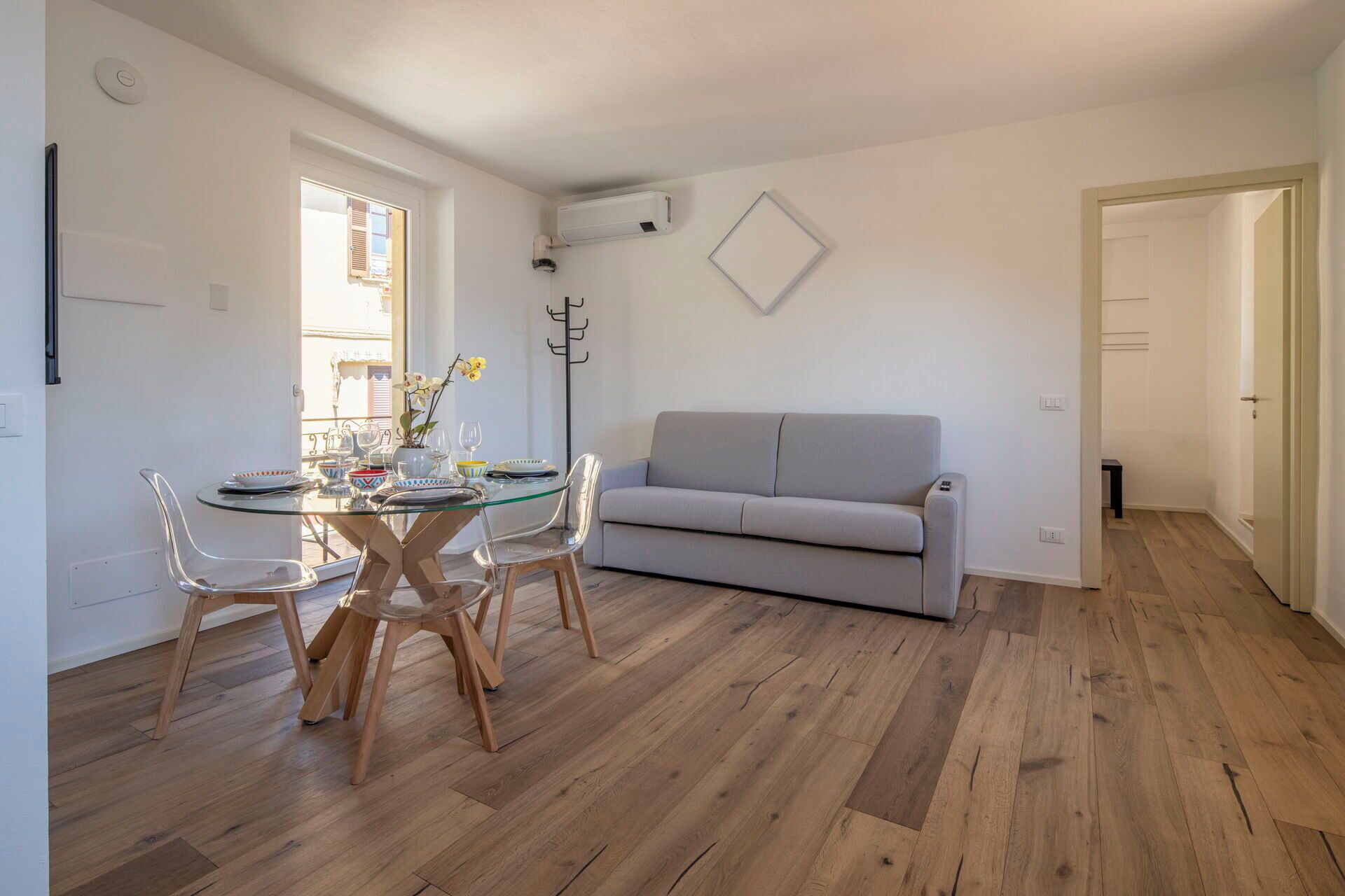 Un comedor con una mesa y sillas de vidrio, un sofá gris y suelo de madera.