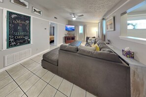 Ein Wohnzimmer mit einem großen, grauen Ecksofa, einem an der Wand befestigten Flachbildfernseher und einem Deckenventilator.