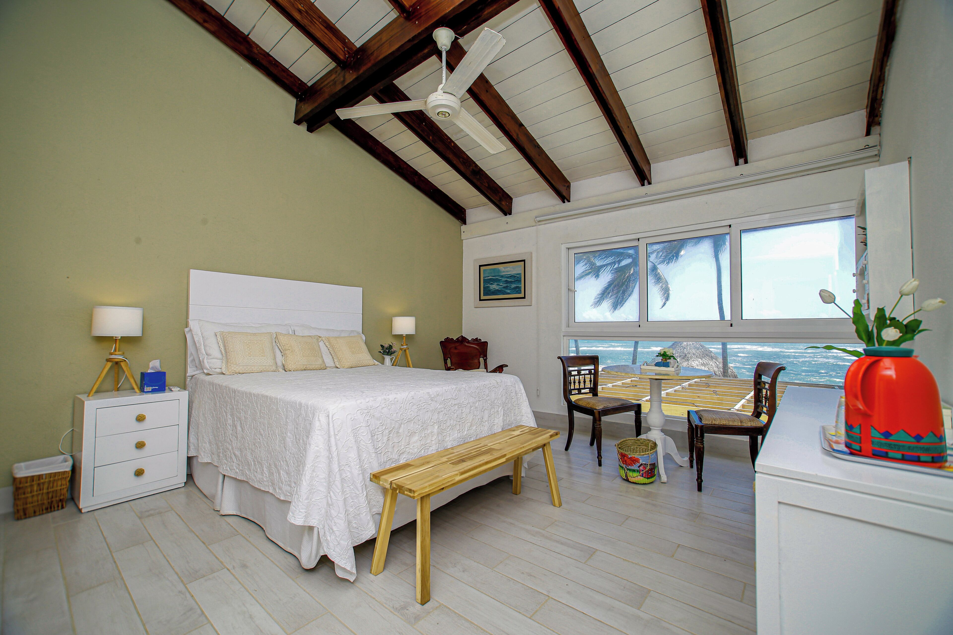 Quarto com cama, mesas de cabeceira, um banco, uma cadeira, uma mesa de jantar e vista para o oceano.