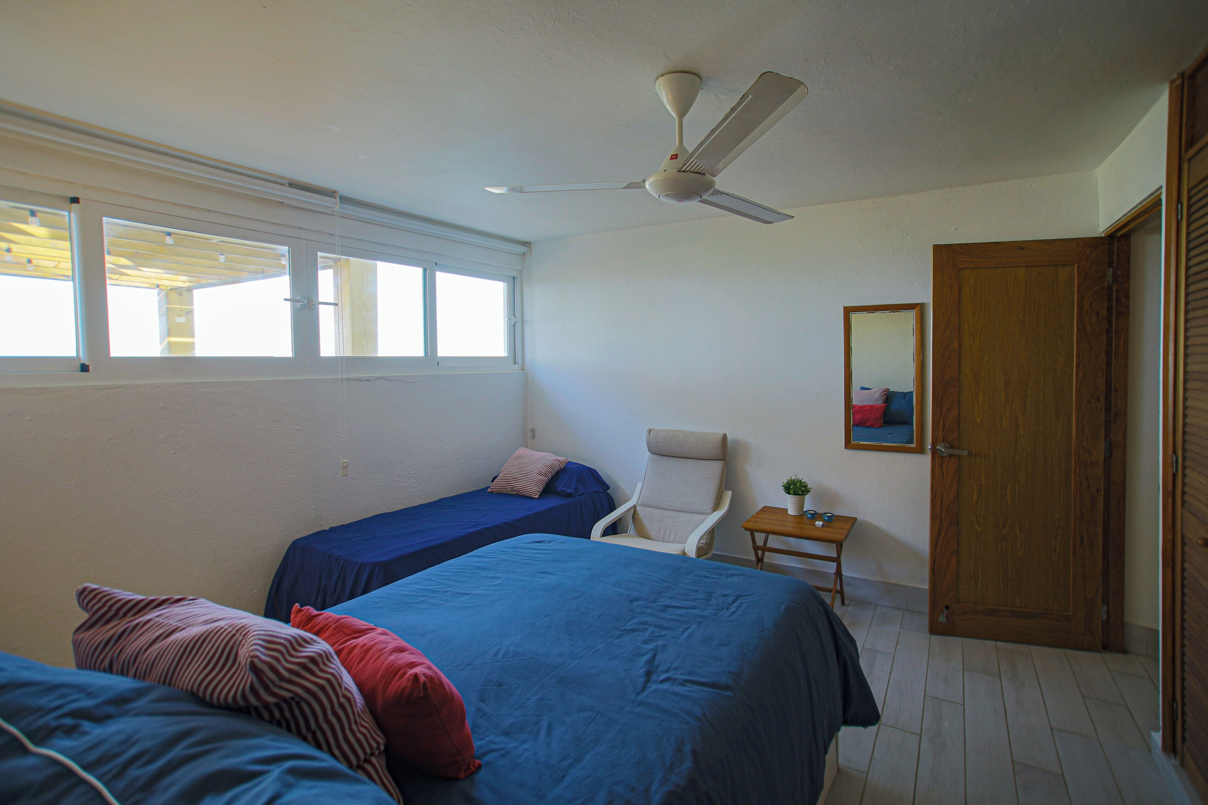 Une chambre avec deux lits, un ventilateur de plafond, une fenêtre, une chaise, une petite table et une porte.