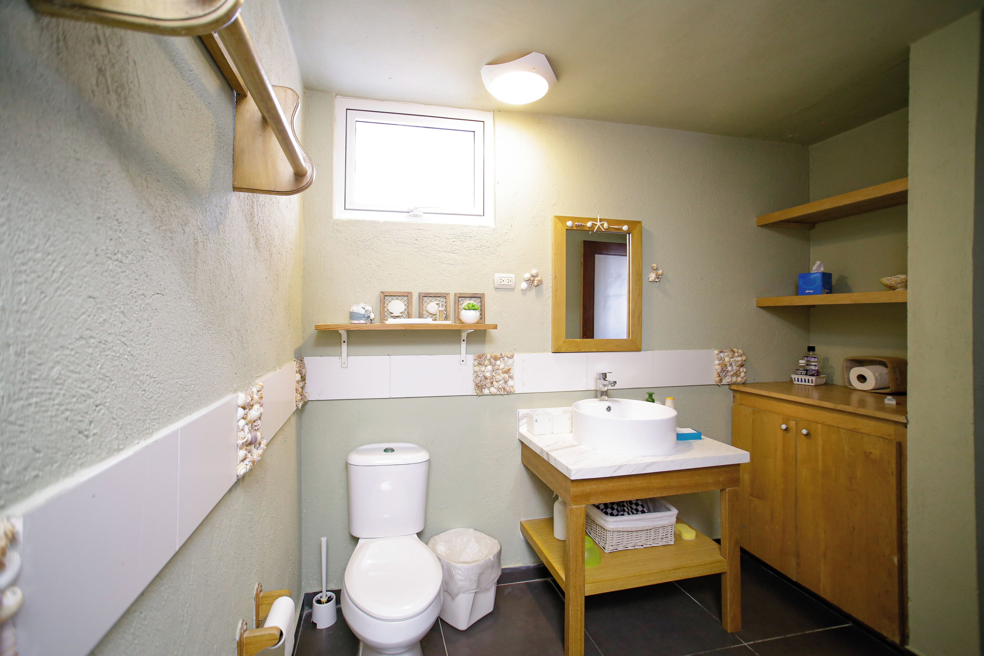 A bathroom with a toilet, sink, and mirror. There is a shelf with toiletries and a small window above the sink.