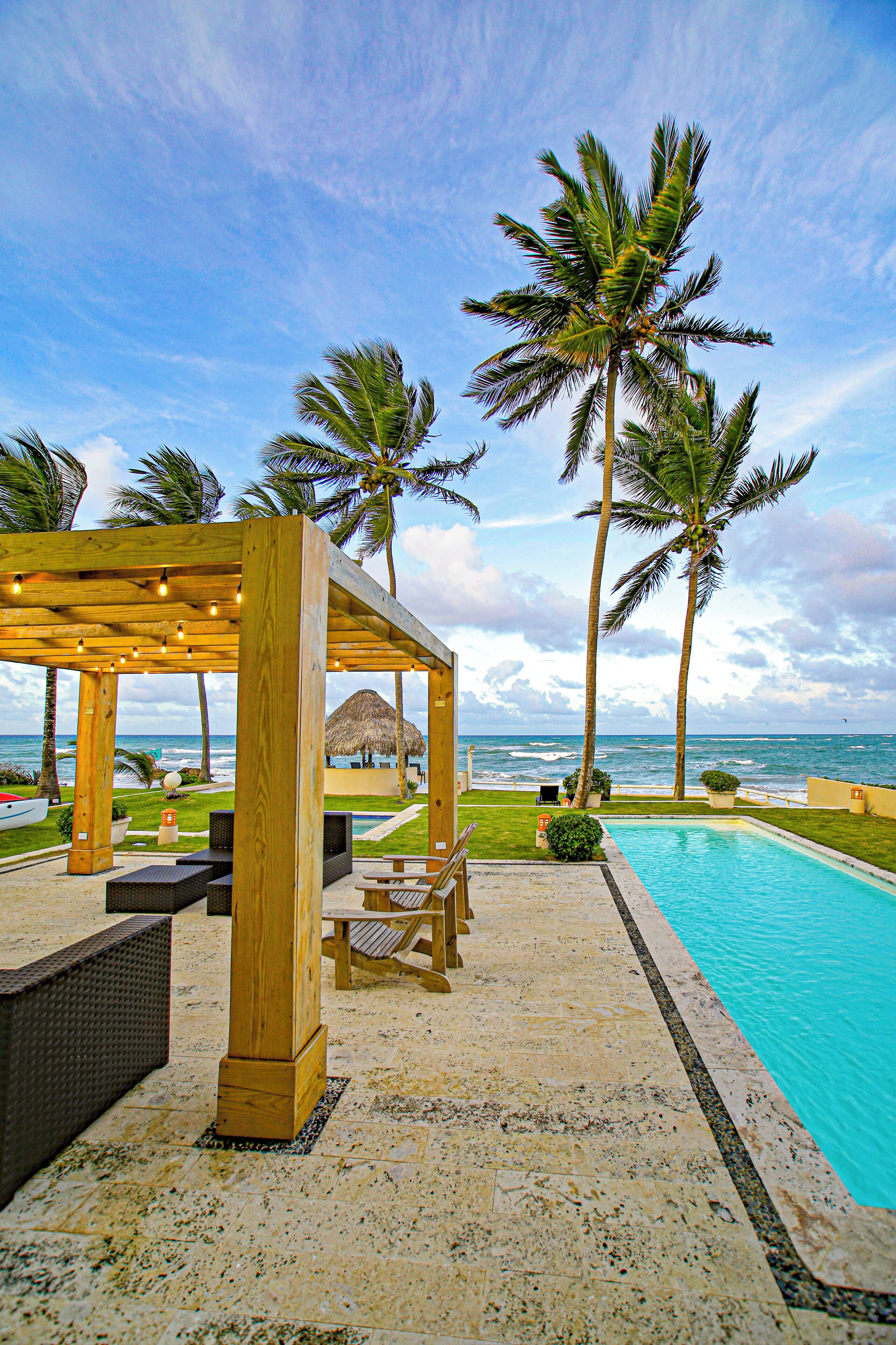 A wooden gazebo with string lights, a pool, palm trees, and a view of the ocean.