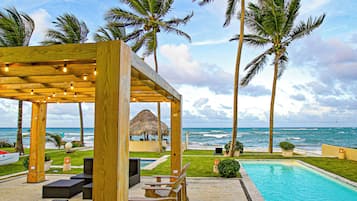A wooden gazebo with string lights, a pool, palm trees, and a view of the ocean.