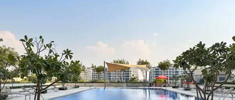 A swimming pool with a tiled deck, surrounded by trees and a building with a red umbrella.