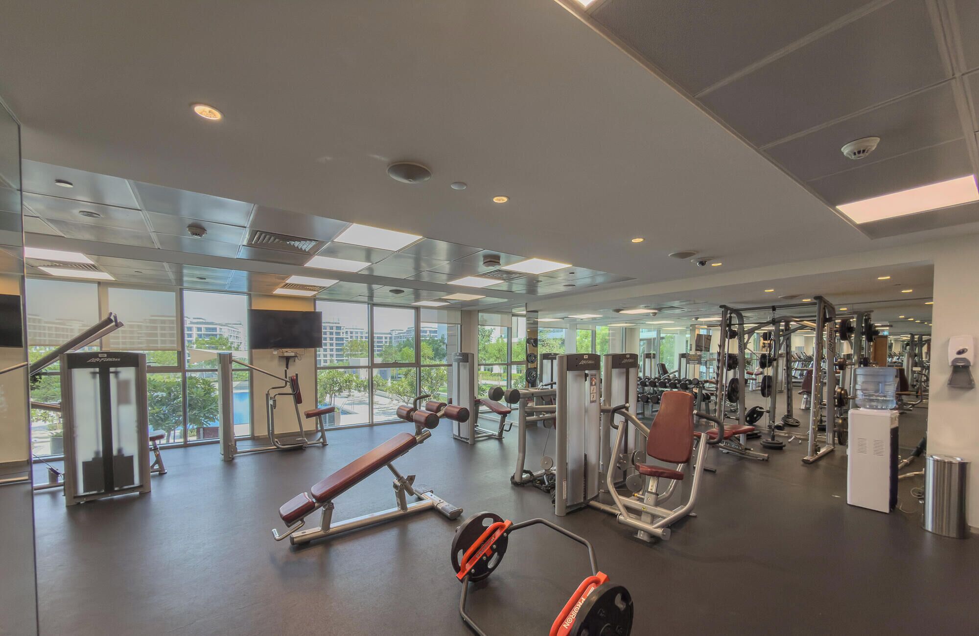 Ett modernt gym med olika typer av träningsutrustning, stora fönster och utsikt över byggnaderna utanför.